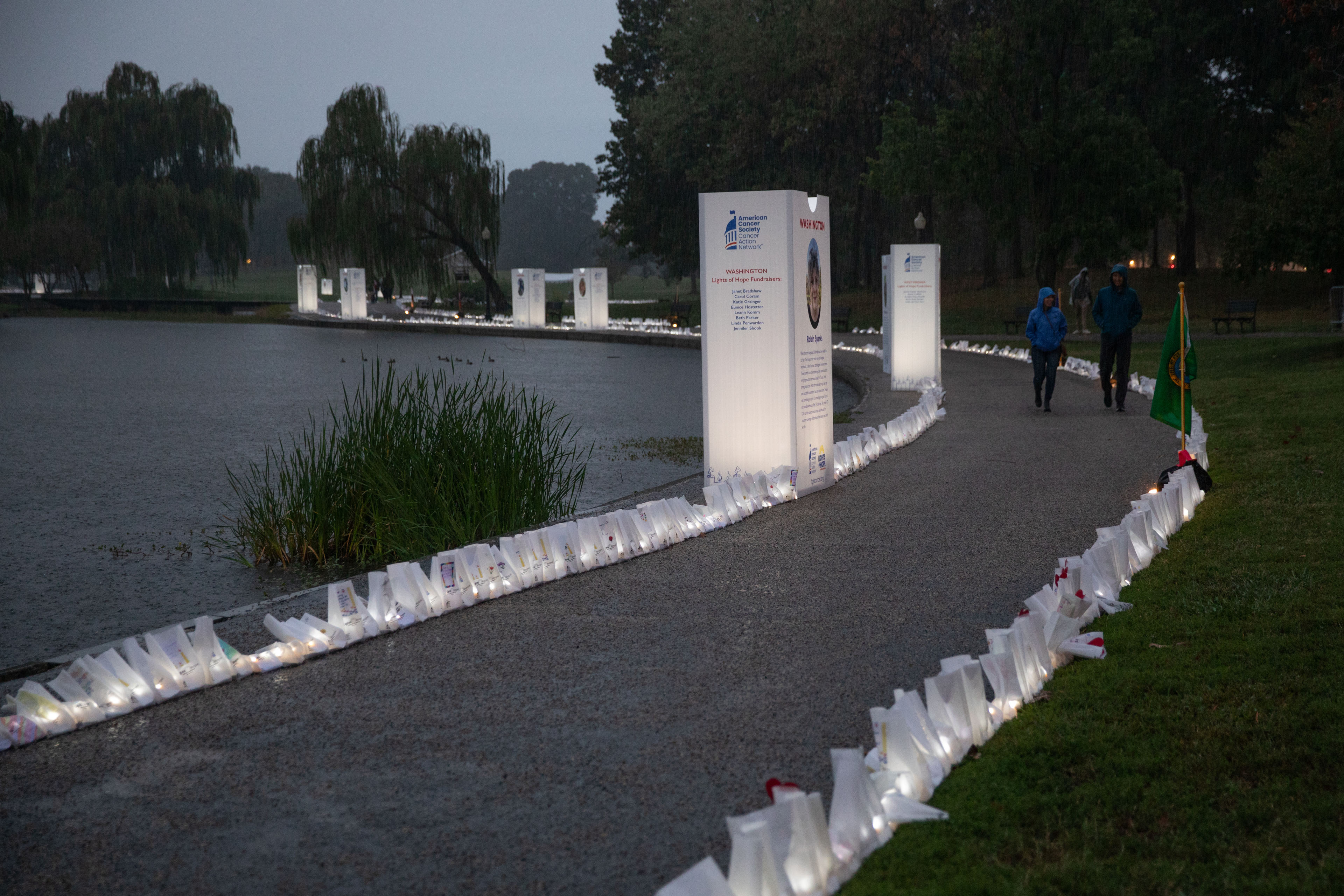 Common-ground-cancer-06 A photograph of a path alongside a body of water. The path is lit with white paper bags that are memorials for people who passed from cancer.